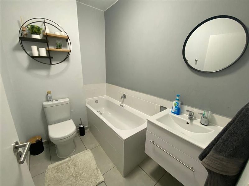 A modern bathroom featuring a built-in bathtub, a white vanity cabinet with a basin, and a toilet, finished with neutral wall tones and tiled flooring.