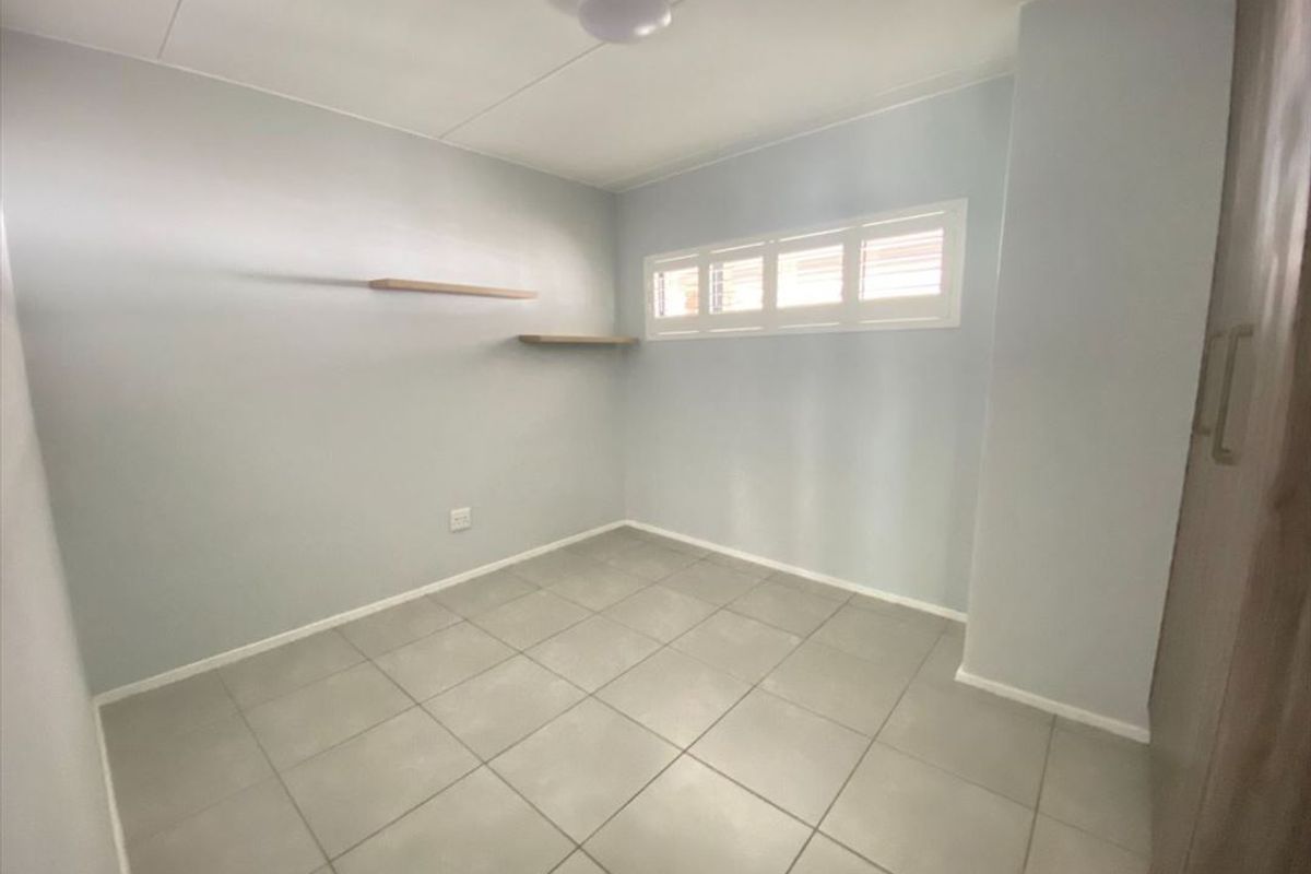 A clean, empty bedroom featuring neutral tiled flooring, built-in cupboards, and floating wall shelves with a high-level louvre window.