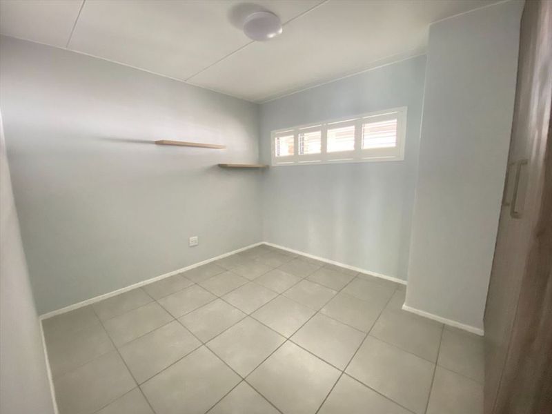 A clean, empty bedroom featuring neutral tiled flooring, built-in cupboards, and floating wall shelves with a high-level louvre window.