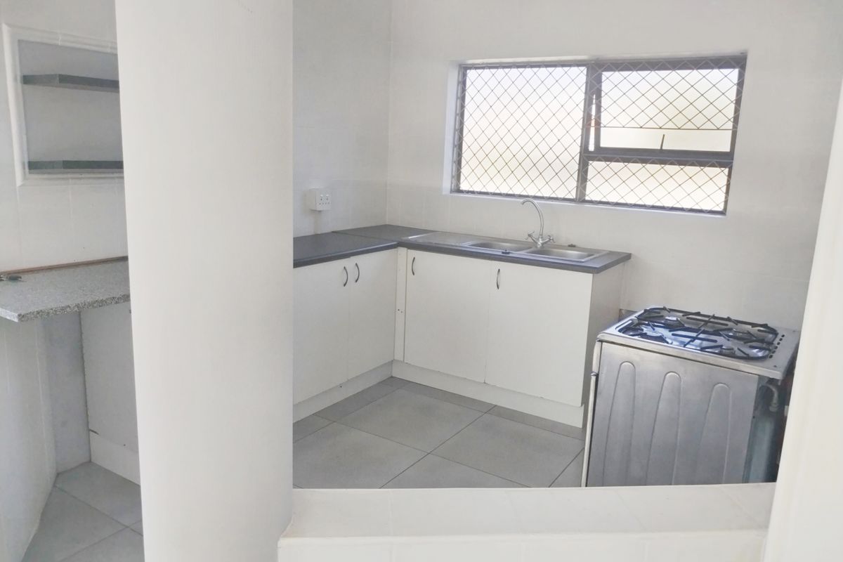 A functional, empty kitchen featuring white cabinetry, laminate countertops, a stainless steel sink, and tiled flooring, with a security-barred window for natural light.