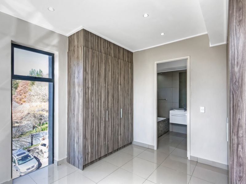 Modern bedroom featuring sleek wood-grain built-in cupboards, neutral tiled flooring, and large windows with views, leading directly into an en-suite bathroom.