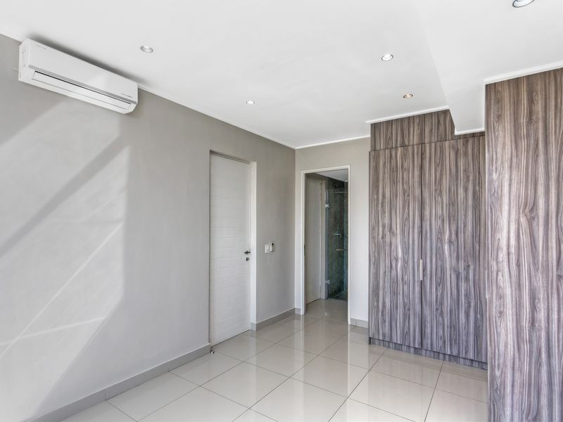 A bright, modern bedroom featuring sleek built-in wood-grain cupboards, neutral tiled flooring, recessed lighting, and a wall-mounted air conditioning unit.