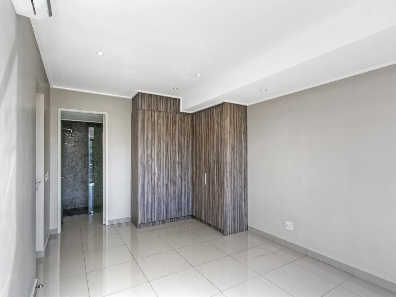A bright, modern bedroom featuring neutral tiled flooring, built-in wood-grain cupboards, recessed lighting, and direct access to an en-suite bathroom.