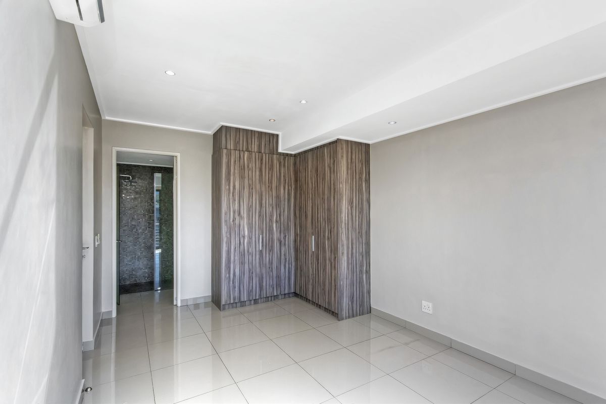 A bright, modern bedroom featuring neutral tiled flooring, built-in wood-grain cupboards, recessed lighting, and direct access to an en-suite bathroom.