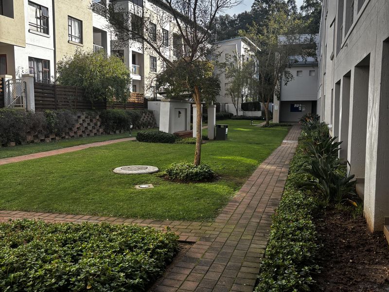 A well-maintained communal garden area between apartment blocks, featuring paved walkways, manicured lawns, and established landscaping.