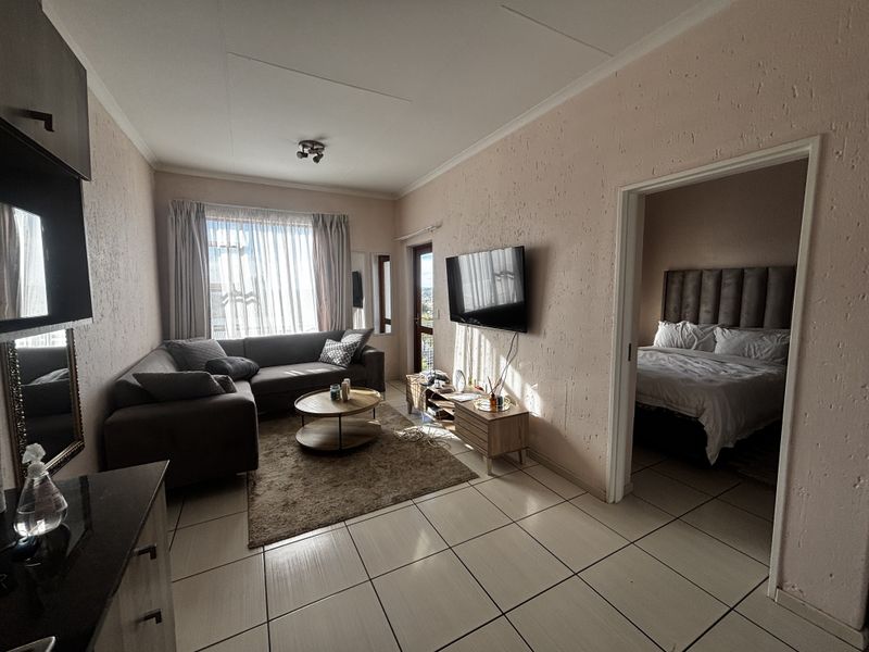 A living room featuring tiled flooring and textured walls, with direct access to an adjacent bedroom and natural light from the balcony door.