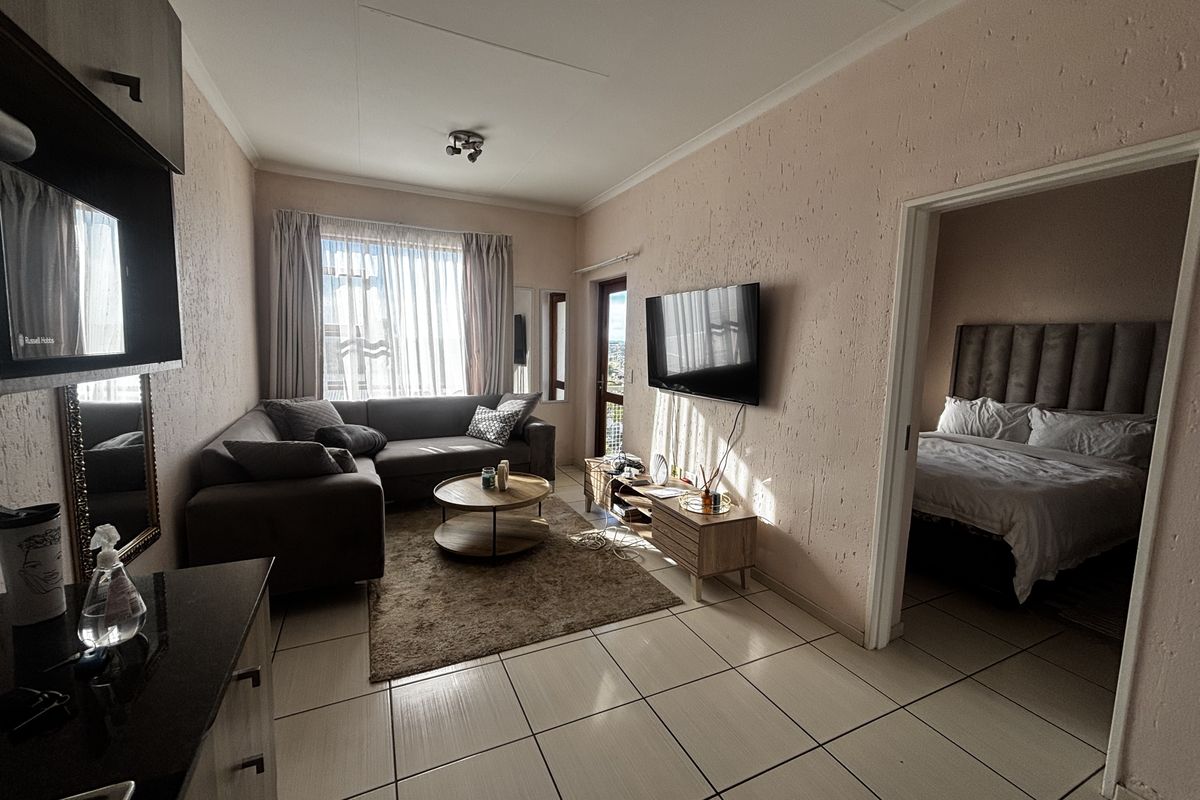A living room featuring tiled flooring and textured walls, with direct access to an adjacent bedroom and natural light from the balcony door.