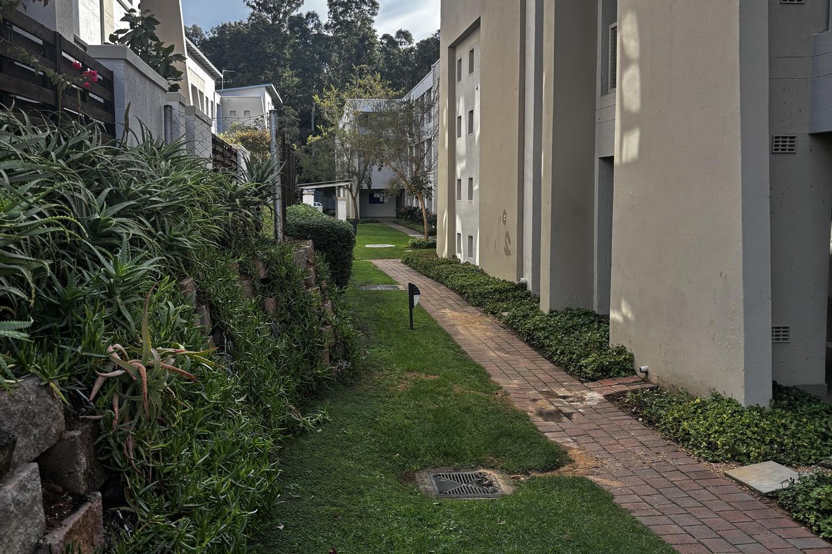A view of the exterior rear grounds featuring a brick paved pathway, landscaped garden beds, and a retaining wall between apartment blocks.