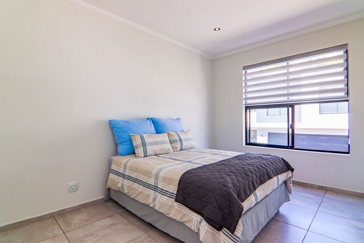 A well-lit bedroom featuring neutral tiled flooring, recessed downlighting, and a large window with modern roller blinds.