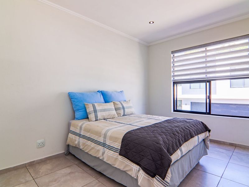 A well-lit bedroom featuring neutral tiled flooring, recessed downlighting, and a large window with modern roller blinds.