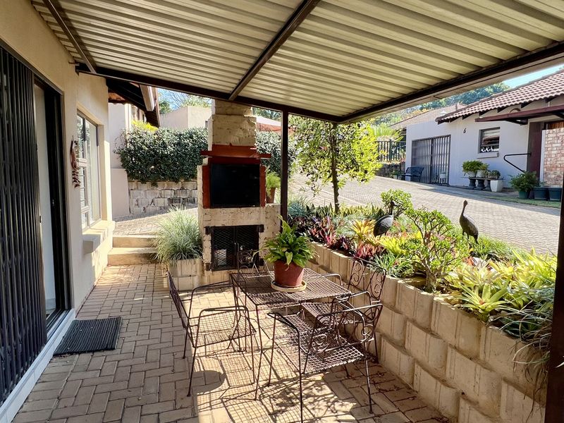 A covered outdoor patio area featuring brick paving, a built-in braai, and a low retaining wall with established garden beds.