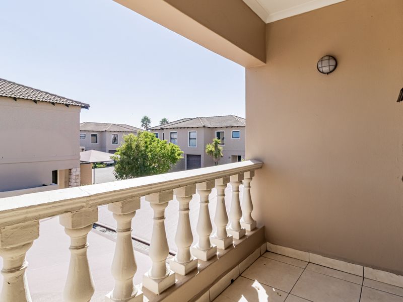 A covered balcony featuring a classic balustrade, tiled flooring, and an exterior wall light, offering views of the surrounding townhouse complex.