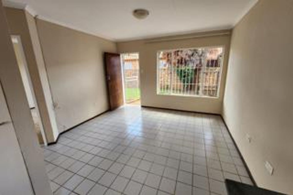 An empty, neutrally painted living room featuring tiled flooring, a large window with security bars, and direct access to the exterior.
