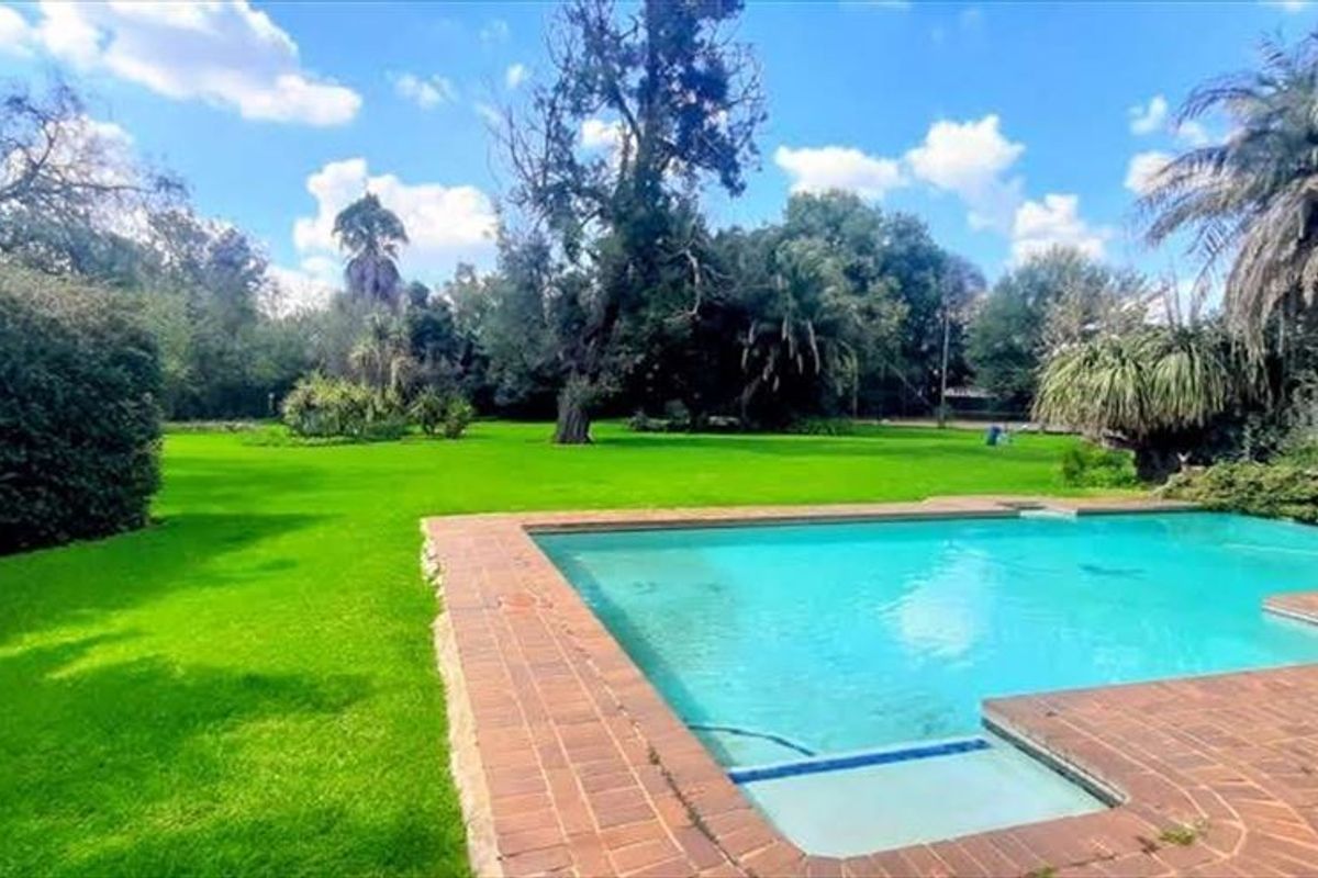 A spacious outdoor pool area featuring a brick-paved surround set within a large, well-maintained garden with mature trees.