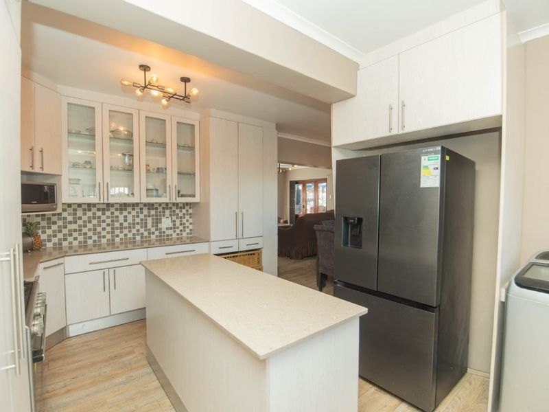 Modern kitchen featuring light-toned cabinetry, stone countertops, a central island, and a mosaic tile backsplash, seamlessly flowing into the adjacent living area.