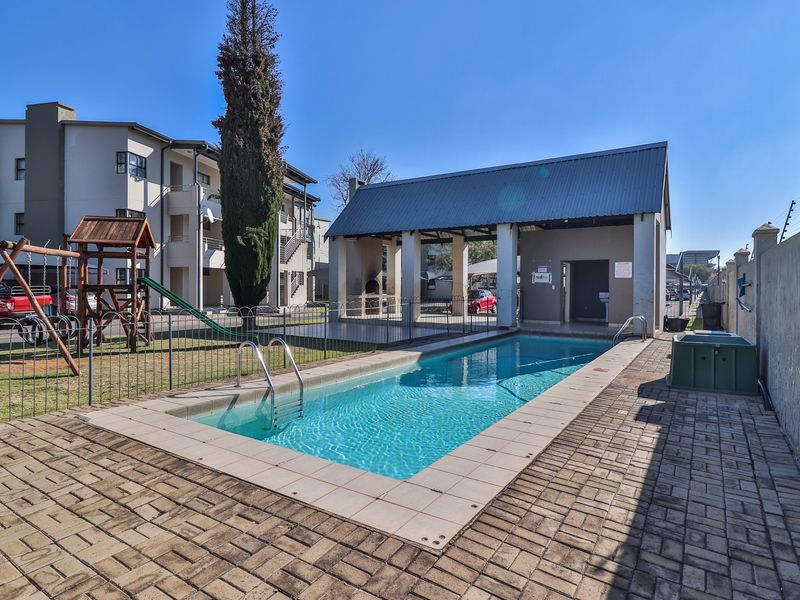 Communal pool area featuring a paved deck, covered patio with built-in braai, and adjacent playground equipment within a secure apartment complex.