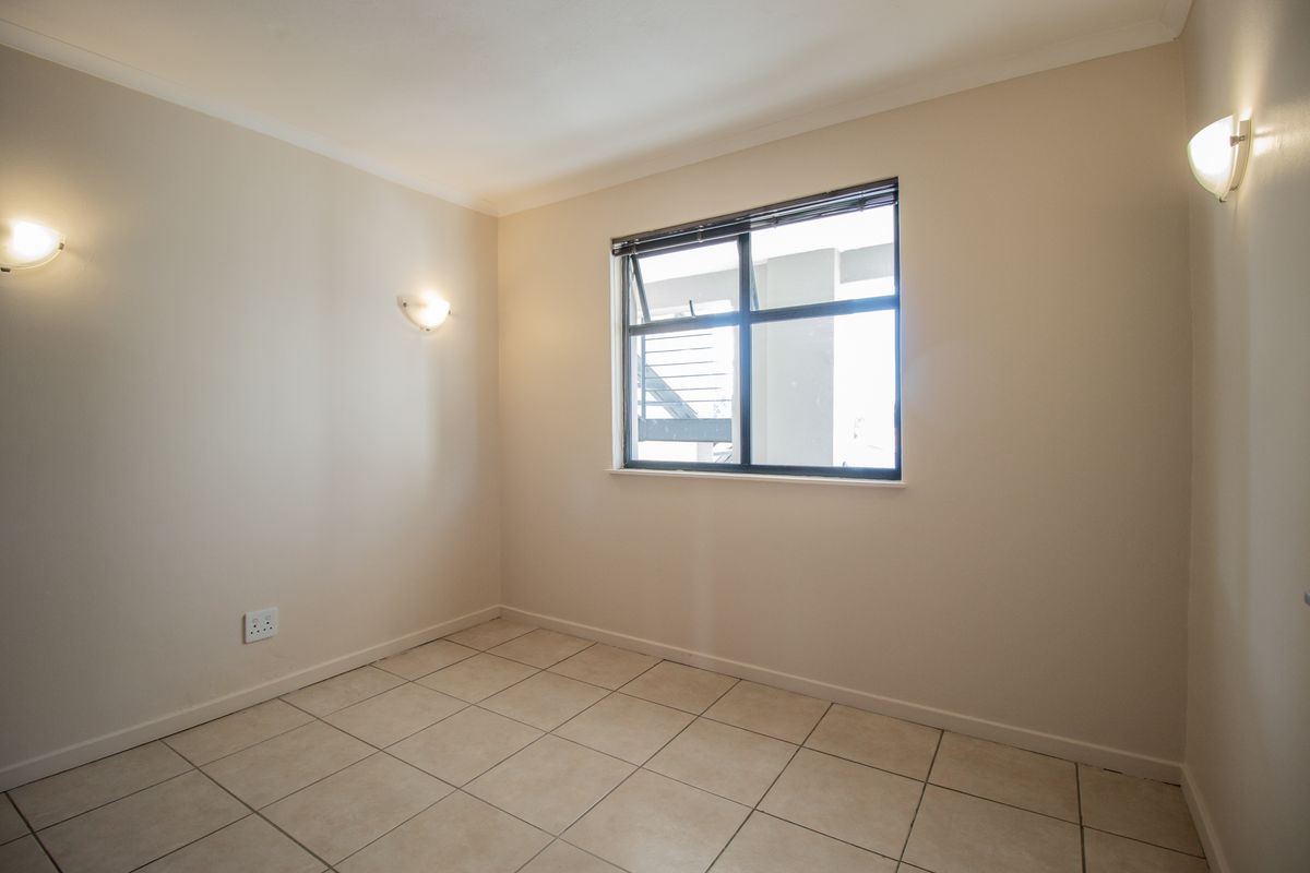 A clean, empty bedroom featuring neutral tiled flooring, wall-mounted lighting, and a window providing natural light.