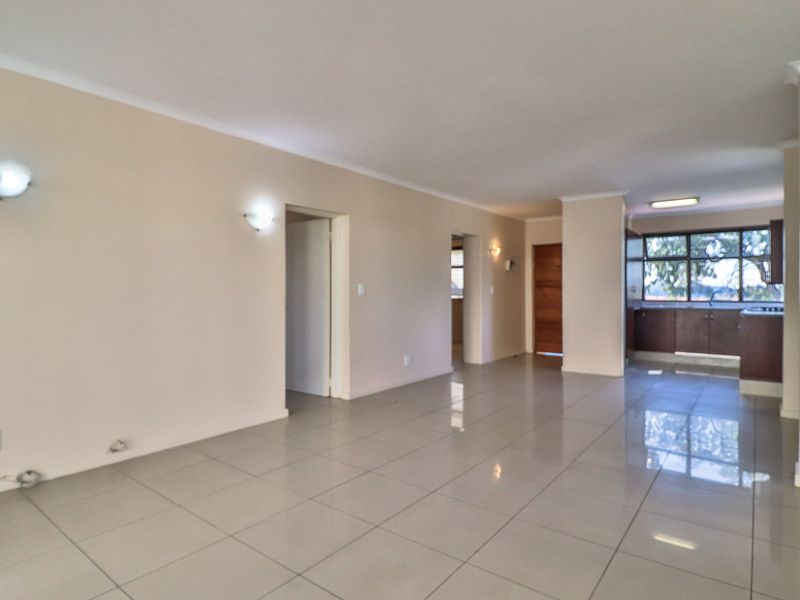 Spacious open-plan living area featuring neutral tiled flooring, wall-mounted lighting, and a functional kitchen space with built-in cabinetry.