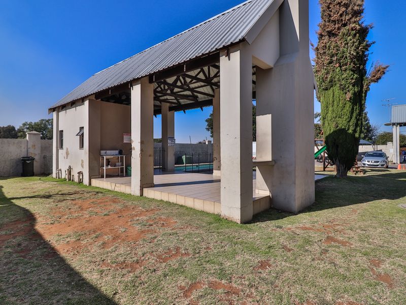 Outdoor communal pool area featuring a covered patio with a built-in braai, set within a landscaped garden space.