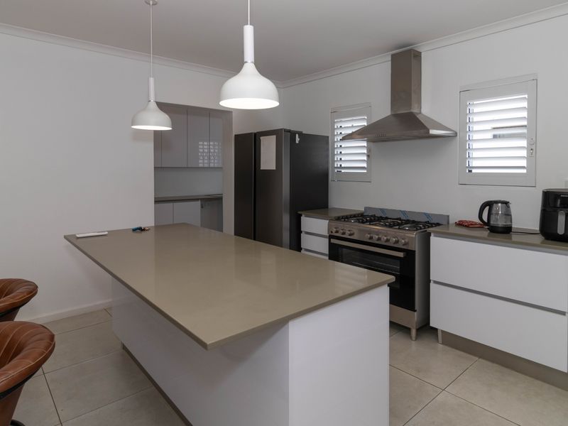 Modern kitchen featuring a central island with stone benchtops, white cabinetry, stainless steel gas stove, and extractor fan, complemented by tiled flooring and plantation shutters.
