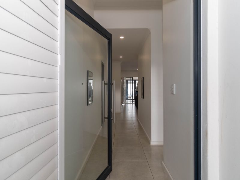 A bright, modern entrance hallway featuring tiled flooring, recessed downlighting, and a contemporary glass pivot door leading into the home.