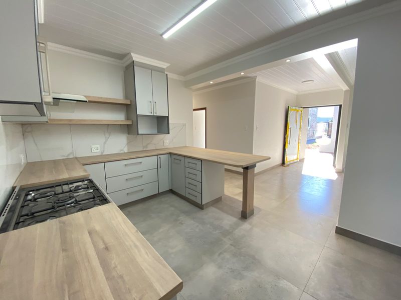 Modern open-plan kitchen featuring light grey cabinetry, timber-look countertops, gas hob, and marble-look splashback, leading to an outdoor entrance.