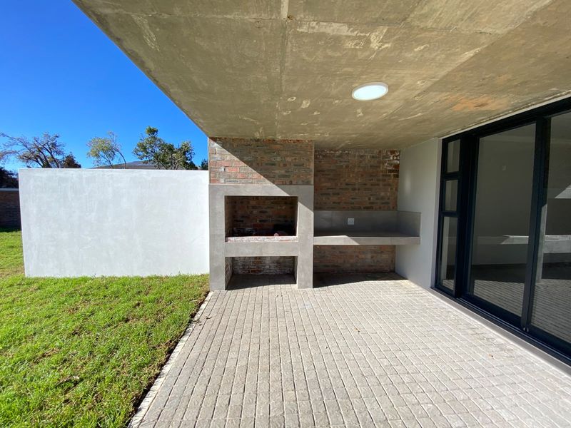A modern covered patio featuring a built-in brick braai, concrete prep counter, and paved flooring, seamlessly connected to the interior via sliding glass doors.