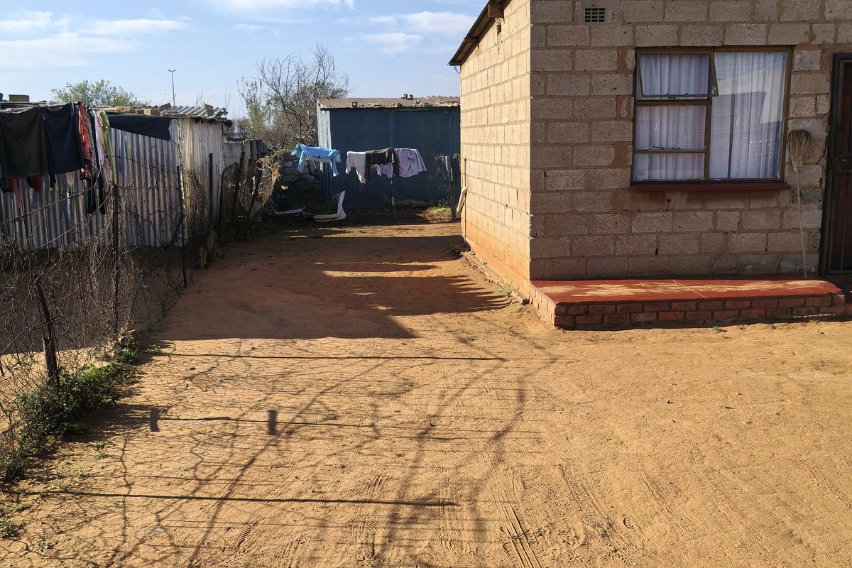 Rear exterior view of a concrete block house featuring a solar water heater and an unpaved yard area.