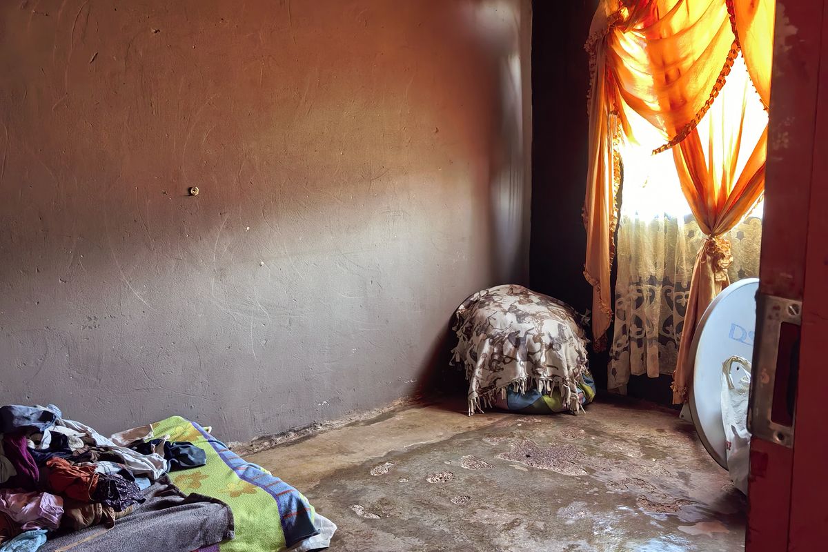 A basic bedroom featuring exposed corrugated metal roofing, plastered walls, and a concrete floor in need of repair.