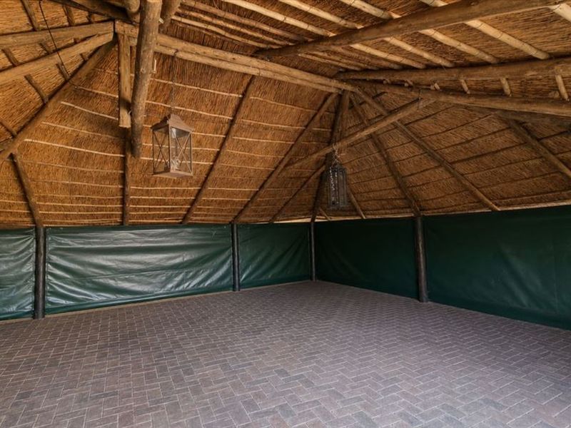 A spacious covered patio area featuring a traditional thatched roof with exposed timber rafters and durable paved flooring, enclosed by protective canvas screens.