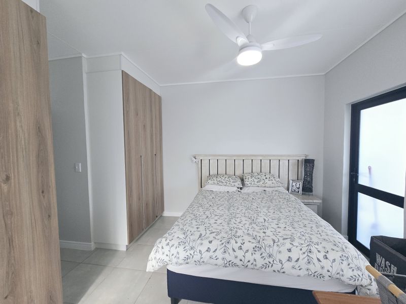 A well-maintained bedroom featuring built-in wood-finish cupboards, a modern ceiling fan, tiled flooring, and a glass panel door providing natural light.