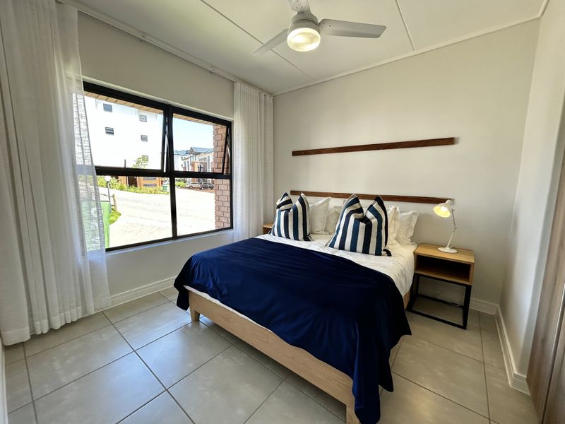 A well-maintained bedroom featuring neutral tiled flooring, a modern ceiling fan, and large aluminium-framed windows allowing for ample natural light.
