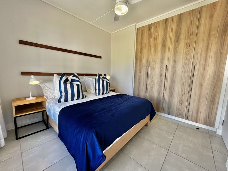 A well-maintained bedroom featuring modern wood-finish built-in cupboards, neutral tiled flooring, and a ceiling fan.