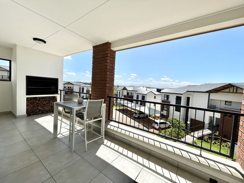 A spacious covered balcony featuring a built-in braai, tiled flooring, and a metal balustrade, offering pleasant views of the surrounding apartment complex.
