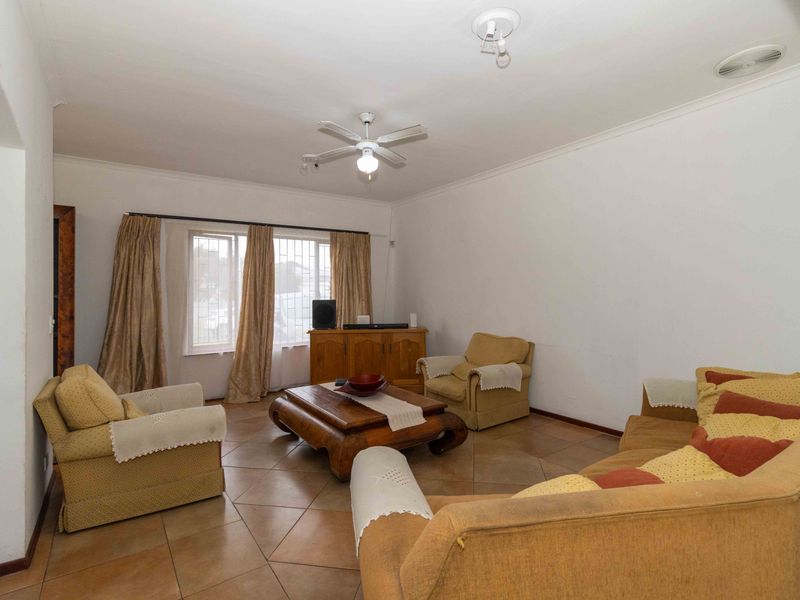 A spacious living room featuring tiled flooring, a ceiling fan, and large windows with security bars, offering a functional and well-lit living space.