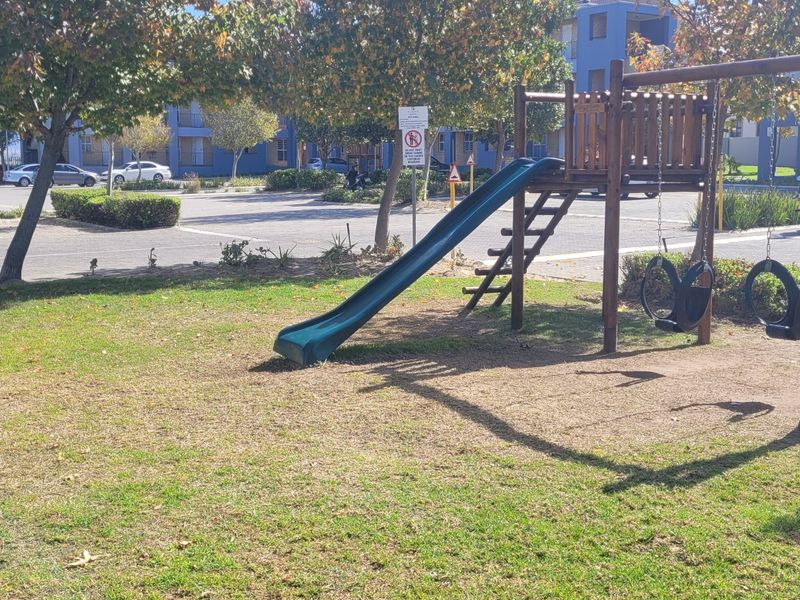 A communal garden area featuring a playground with a slide and swings, situated adjacent to an apartment complex parking lot.