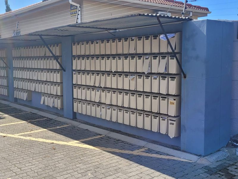 Exterior view of a large communal mailbox bank with protective metal awning, situated along the perimeter wall of the apartment complex.