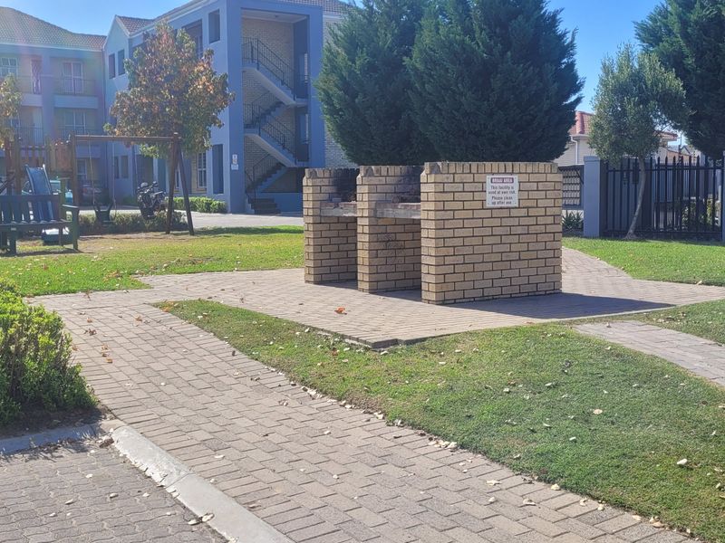 Communal garden area featuring a brick built-in braai facility, paved pathways, and well-maintained lawn within the apartment complex grounds.
