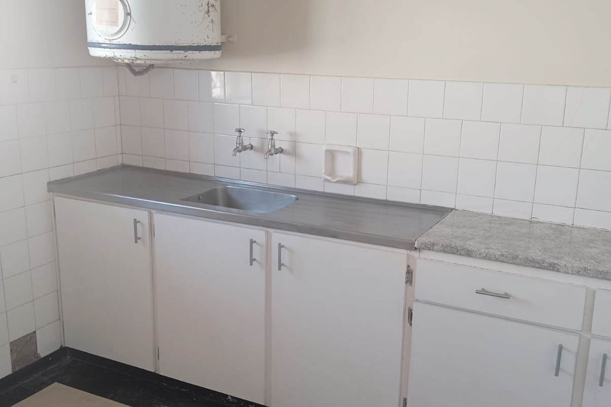 A dated kitchen area featuring basic white cabinetry, a stainless steel sink, tiled splashbacks, and a wall-mounted electric water heater.