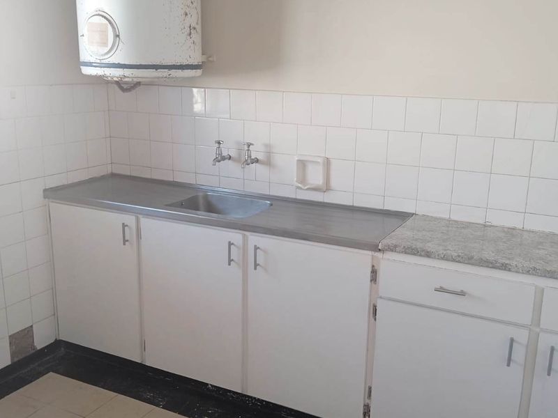 A dated kitchen area featuring basic white cabinetry, a stainless steel sink, tiled splashbacks, and a wall-mounted electric water heater.