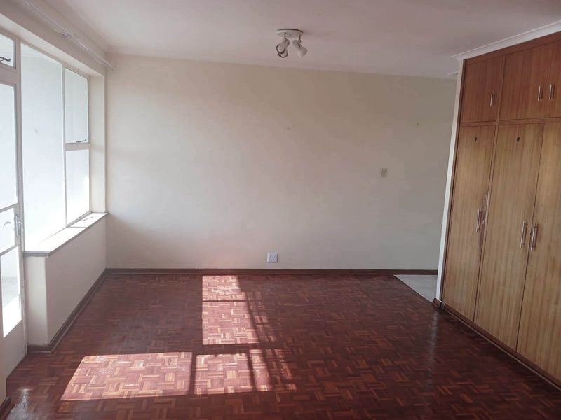 A spacious, empty bedroom featuring classic parquet flooring, large windows for natural light, and extensive built-in wooden cabinetry for storage.