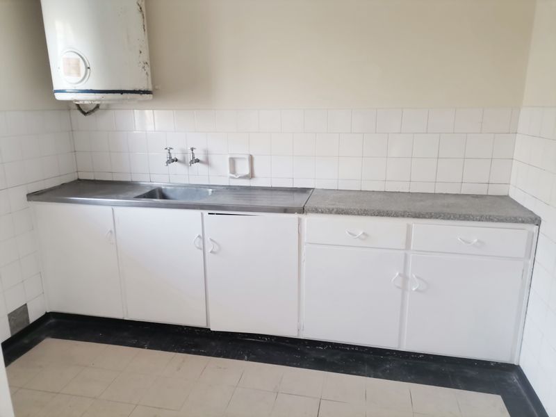 A dated kitchen area featuring white cabinetry, a stainless steel sink, tiled splashbacks, and a wall-mounted electric water heater.