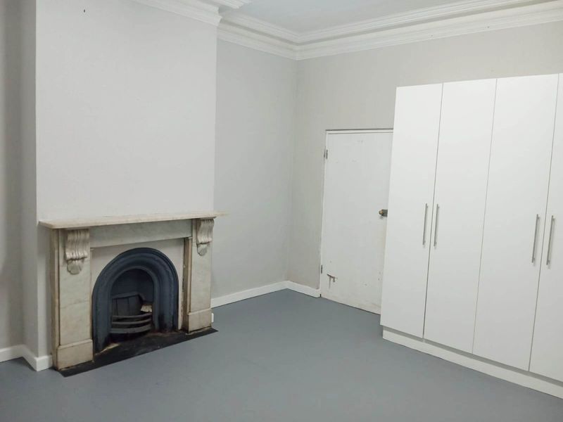 A bright, empty bedroom featuring a classic fireplace with a mantelpiece, built-in white wardrobes, and neutral grey flooring.