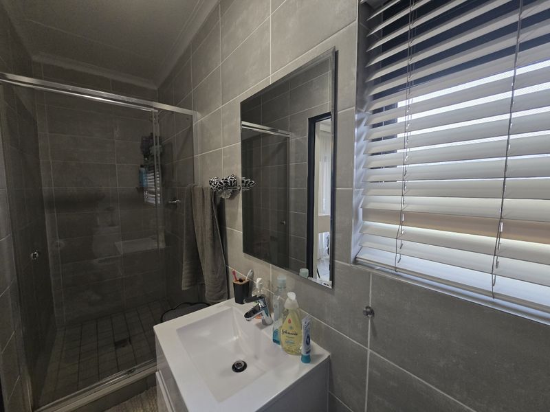Modern bathroom featuring a glass shower enclosure, vanity basin with mixer tap, and neutral grey wall tiling with venetian blinds.