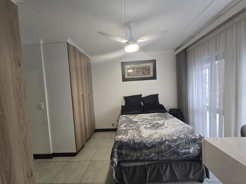 A well-maintained bedroom featuring built-in wooden cupboards, tiled flooring, a ceiling fan, and direct access to a balcony through sliding glass doors.