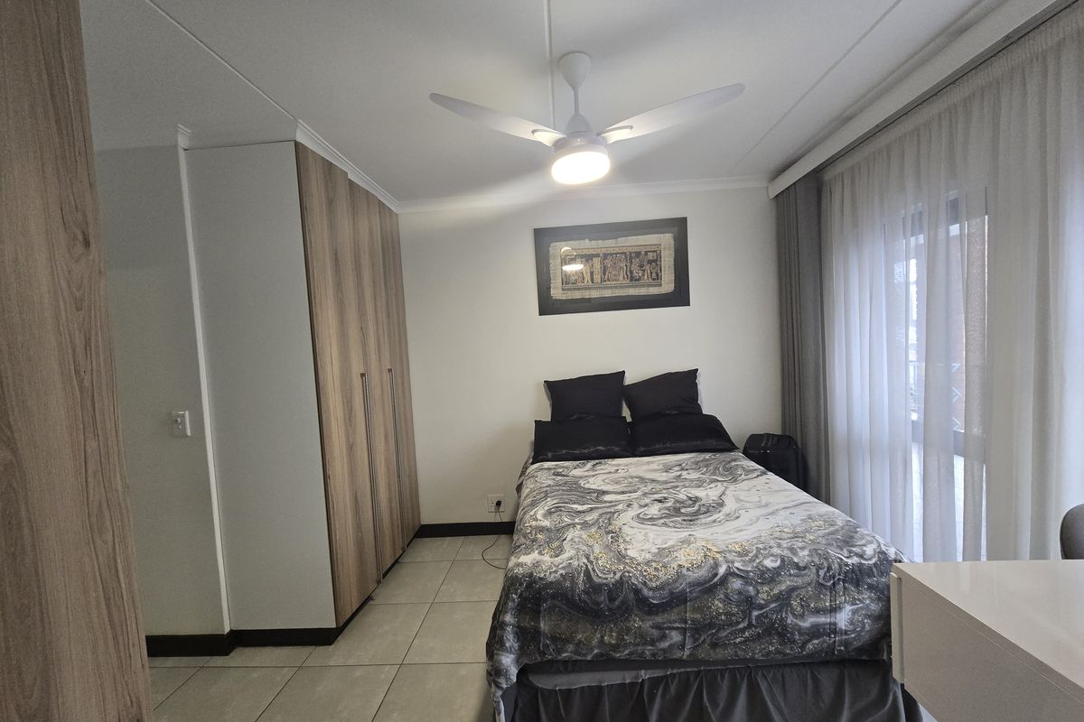 A well-maintained bedroom featuring built-in wooden cupboards, tiled flooring, a ceiling fan, and direct access to a balcony through sliding glass doors.