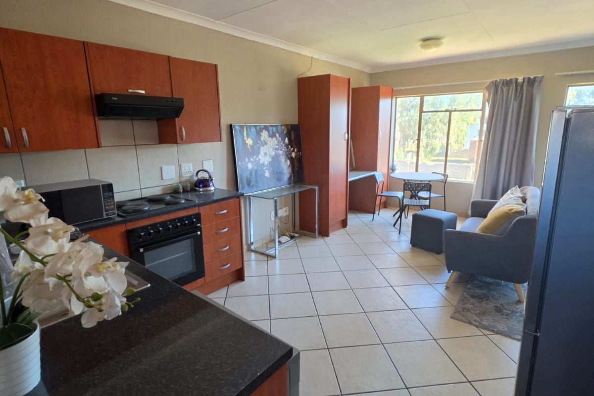 Open-plan kitchen and living area featuring tiled flooring, wooden cabinetry, and granite countertops, with natural light from a large window.