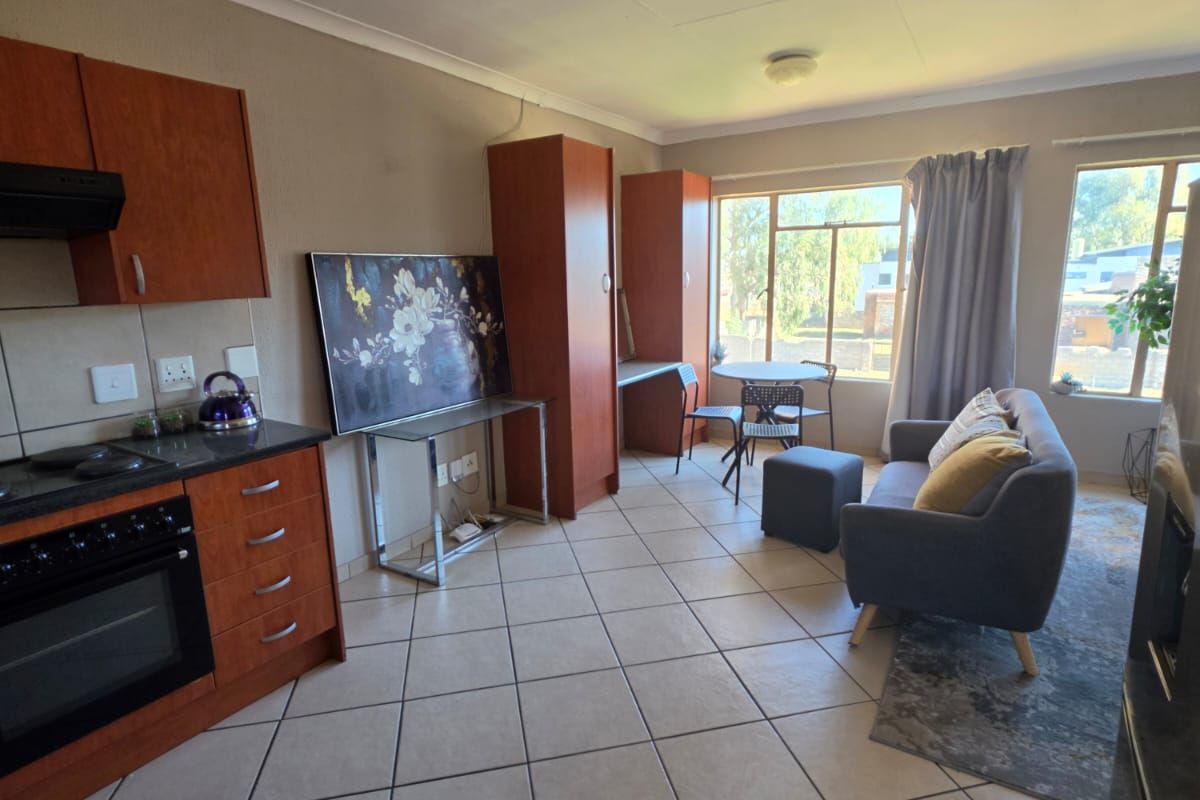 Open-plan living area featuring a functional kitchen with built-in cabinetry, electric oven, and hob, seamlessly flowing into a bright lounge space with tiled flooring.