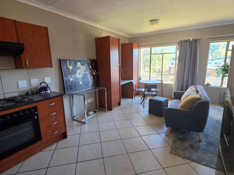 Open-plan living area featuring a functional kitchen with built-in cabinetry, electric oven, and hob, seamlessly flowing into a bright lounge space with tiled flooring.