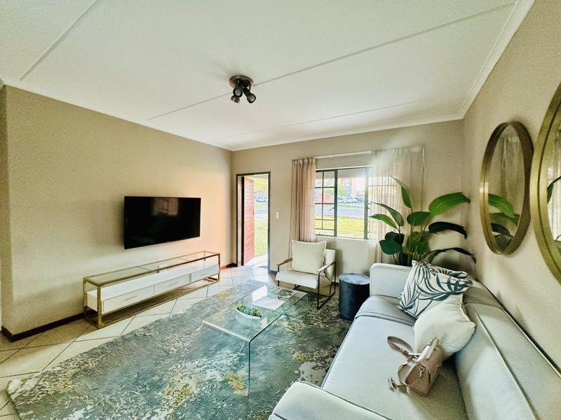 A well-maintained living room featuring tiled flooring, neutral wall tones, and a window with security bars, providing a bright and functional living space.